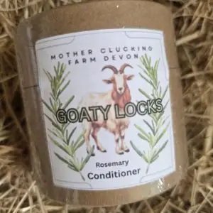Close-up of a brown bottle with a label reading 'Goaty Locks' and shows a goat illustration surrounded by rosemary sprigs, resting in hay. Scrolling text includes 'Rosemary Conditioning' (brand: Mother Clucking Farm Devon).